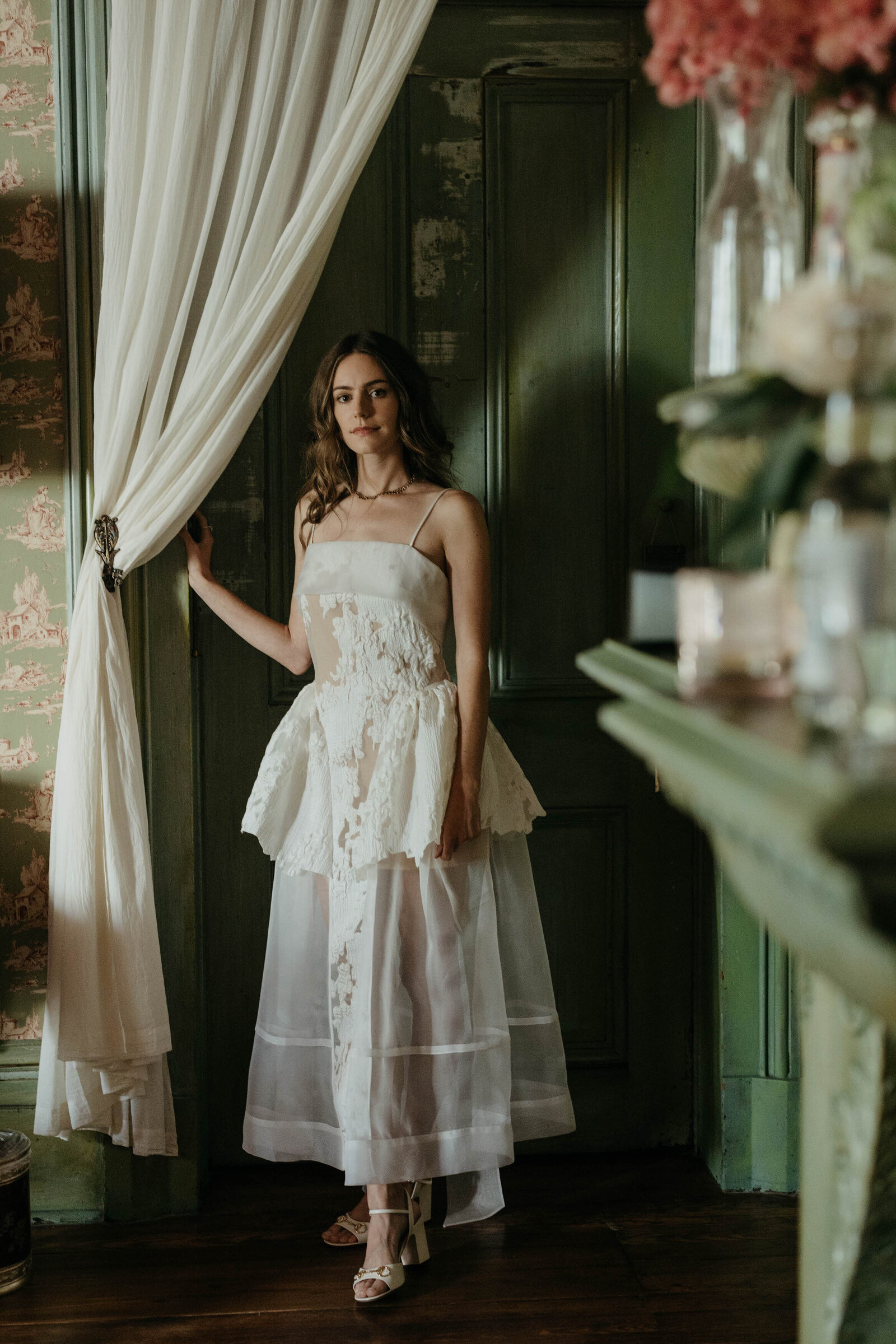 Bride in a white lace gown beside a sheer curtain and green panelled door